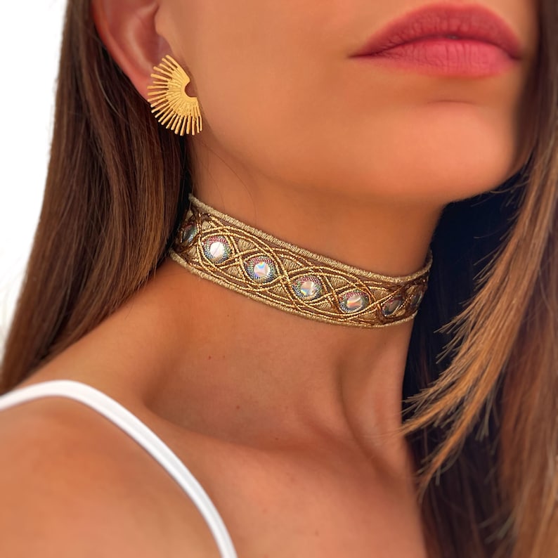 May include: A gold-colored choker necklace with iridescent stones and a matching gold earring. The choker features an intricate woven design with oval-shaped iridescent stones. The earring is a half-circle shape with vertical lines.
