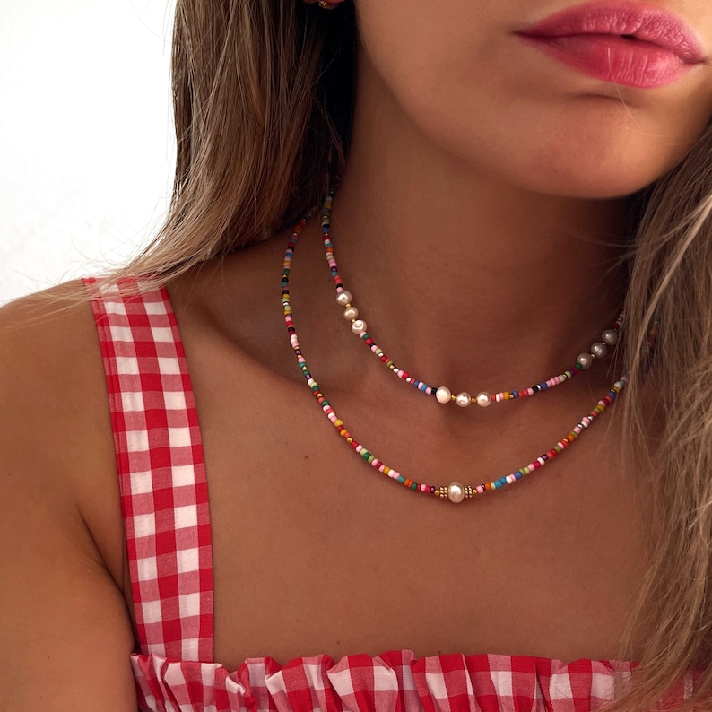 May include: A layered necklace with colorful beads and white pearls. The necklace is worn by a person with long brown hair and a red and white gingham top.