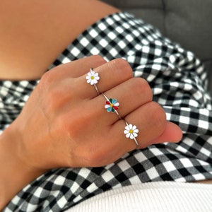 May include: Three silver rings featuring daisy flower designs. The middle ring has a colourful daisy with red, yellow, green, and blue petals. The other two rings have white daisies with yellow centres.