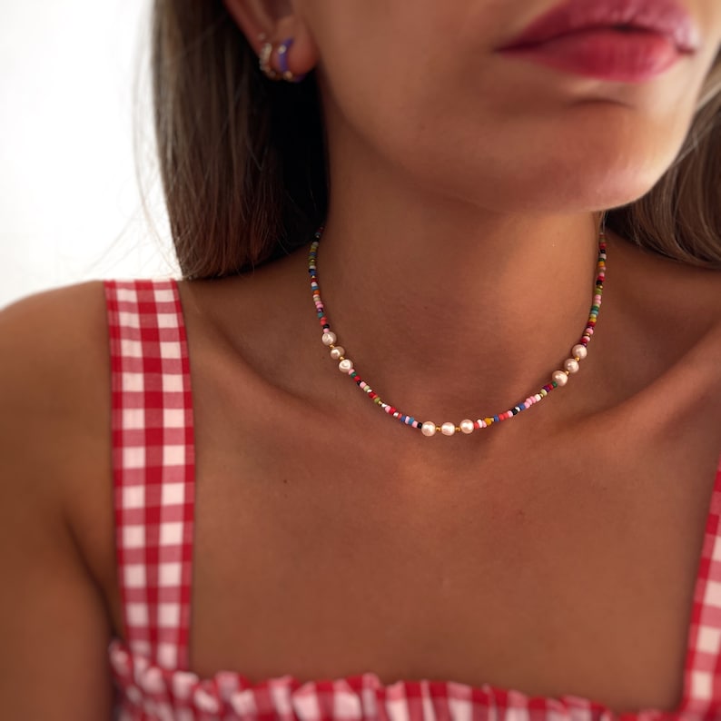 May include: A colorful beaded necklace with small white pearls. The necklace is on a person wearing a red and white gingham top.