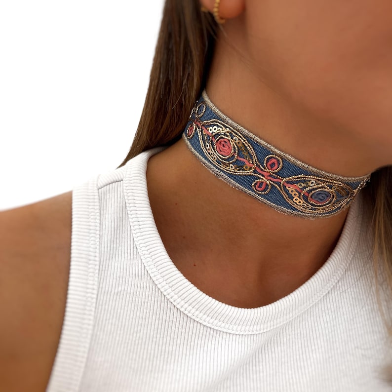 May include: A blue choker necklace with gold and pink embroidery. The choker is made of a fabric with a woven texture.