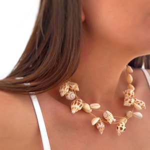 Puede incluir: Un collar que presenta múltiples conchas marinas en espiral con patrones en tonos bronce y blanco, junto con conchas cauri lisas de color crema, ensartadas en un hilo metálico fino de color dorado, creando un diseño delicado y superpuesto.