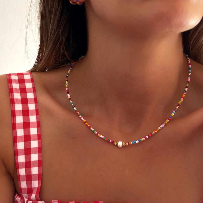 May include: A colorful beaded necklace with a single pearl accent. The necklace is made of small, multi-colored beads and has a gold clasp.