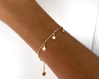 Puede incluir: Un brazalete de cadena dorada con pequeños colgantes en forma de estrella. El brazalete está en la muñeca de una persona.