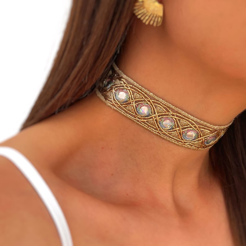 May include: Gold beaded choker necklace with iridescent stones. The necklace features a woven design with a metallic gold finish and is worn around the neck. A gold-colored earring is visible in the background.