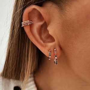 Puede incluir: Puño de oreja y pendientes de aro dorados. El puño de oreja presenta tres bandas apiladas con piedras preciosas de colores. Los pendientes de aro están adornados con una fila de gemas vibrantes y multicolores. Las joyas se muestran en una oreja.