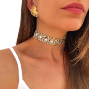 May include: Gold-toned choker necklace with a woven design and clear, round embellishments. The necklace is paired with matching gold sunburst earrings. The jewelry is worn on a person with fair skin and long brown hair.