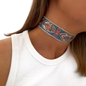 May include: A blue choker necklace with gold and pink embroidery and sequins. The choker is made of a soft fabric and has a delicate design.