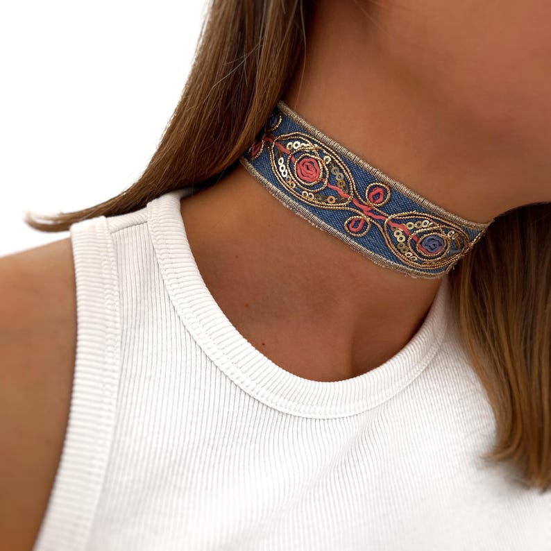 May include: A blue choker necklace with gold and pink embroidery and sequins. The choker is made of a soft fabric and has a delicate design.