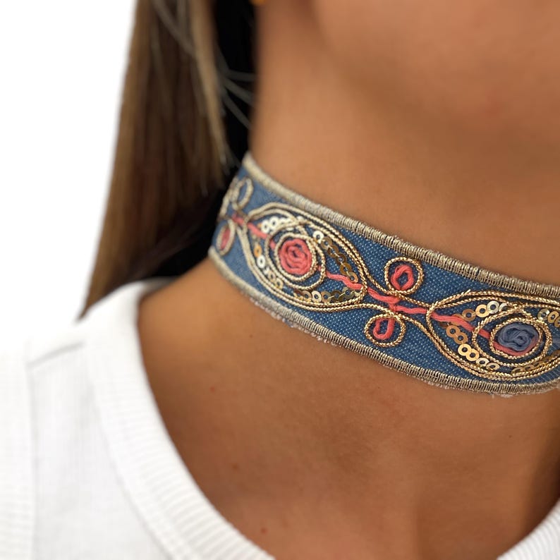 May include: A blue denim choker necklace with gold and pink embroidery. The choker features a repeating pattern of stylized flowers and sequins.