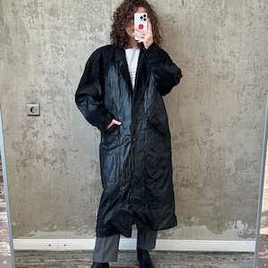 Puede incluir: Una larga gabardina negra con cierre de cremallera y cuello alto. La gabardina está hecha de una tela brillante acolchada.
