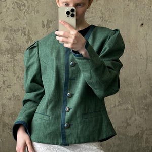 May include: A green jacket with dark blue trim and silver buttons. The jacket has a tailored fit with a round neckline and puffed sleeves. The person in the image is holding a phone in front of their face, obscuring their features. The bottom half of a white lace skirt is visible.