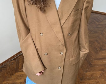 Könnte beinhalten: Ein brauner zweireihiger Blazer mit einer langen, übergroßen Passform. Der Blazer hat vier goldene Knöpfe und ist aus einem glatten, strukturierten Stoff gefertigt.