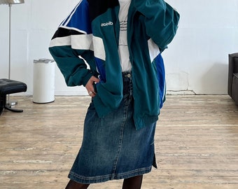 Puede incluir: Una chaqueta azul turquesa con bloques de color negro, blanco y azul. La chaqueta tiene una cremallera y un logotipo que dice "adidas" en el lado izquierdo del pecho. La chaqueta se lleva con una camiseta blanca y una falda vaquera azul.