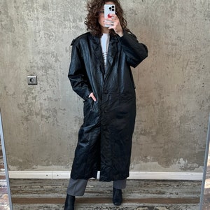 Puede incluir: Una larga gabardina de cuero negro con un cierre de botón único y bolsillos. La gabardina la lleva puesta una persona con el pelo rizado.