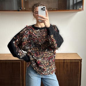 Puede incluir: Un suéter texturizado multicolor con mangas negras y cuello redondo. El suéter presenta una mezcla de hilos rojos, marrones y crema, creando un diseño único. Las mangas son de color negro liso, y el estilo general es casual y cómodo.