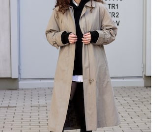 Può includere: Un trench beige con un maglione nero con cappuccio sotto. Il cappotto ha una chiusura a zip e una cintura. La persona indossa collant neri e stivali neri.