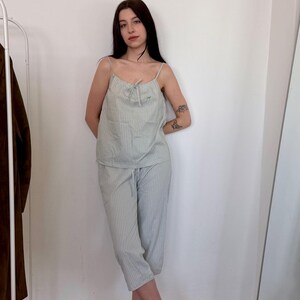 May include: A light green striped pajama set. The set includes a camisole top with thin straps and matching cropped pants. The outfit is made of a lightweight fabric. The model is wearing green mules. The background is a white wall and a wooden floor.