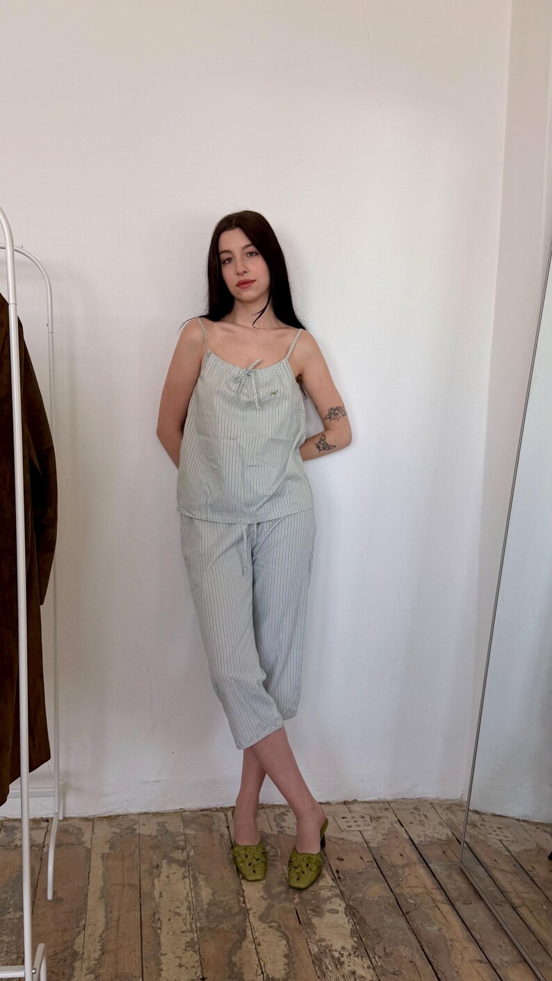 May include: A light green striped pajama set. The set includes a camisole top with thin straps and matching cropped pants. The outfit is made of a lightweight fabric. The model is wearing green mules. The background is a white wall and a wooden floor.