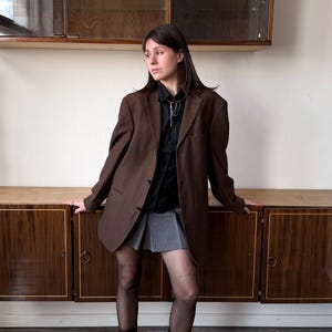 Op de afbeelding: Een bruine blazer gecombineerd met een zwart overhemd, een grijze plooirok en bordeauxrode laarzen. De outfit wordt gecompleteerd met doorschijnende zwarte panty's. De achtergrond toont een houten kast en een witte muur.
