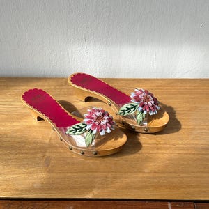 Op de afbeelding: Een paar houten klompen met lage hakken, voorzien van een doorzichtige band versierd met een driedimensionale stoffen bloem in roze, witte en groene tinten. Het voetbed is bekleed met felmagenta met een geschulpte rand en kleine metalen studs.