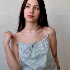 May include: Light blue and white striped camisole top with thin straps and a small green embroidered design. The top is part of a matching pajama set. The fabric has vertical stripes.