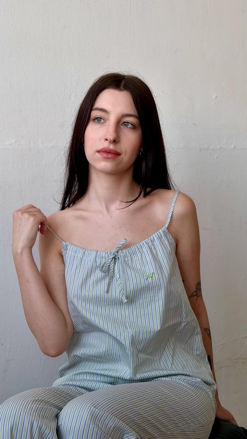 May include: Light blue and white striped camisole top with thin straps and a small green embroidered design. The top is part of a matching pajama set. The fabric has vertical stripes.