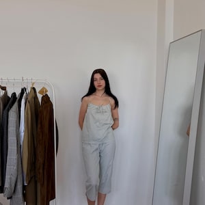May include: A light blue striped pajama set, consisting of a camisole top and cropped pants. The top has a bow detail at the neckline. A clothing rack with various jackets and a full-length mirror are in the background.