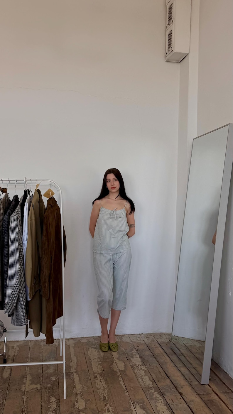 May include: A light blue striped pajama set, consisting of a camisole top and cropped pants. The top has a bow detail at the neckline. A clothing rack with various jackets and a full-length mirror are in the background.