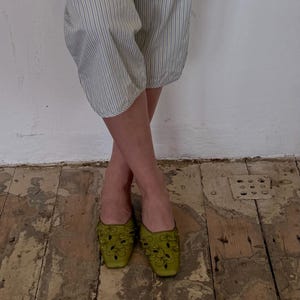 May include: A pair of olive green, embellished mules with a square toe. The shoes are adorned with dark green floral designs and small, round embellishments. The person is wearing striped pants with blue and white stripes.