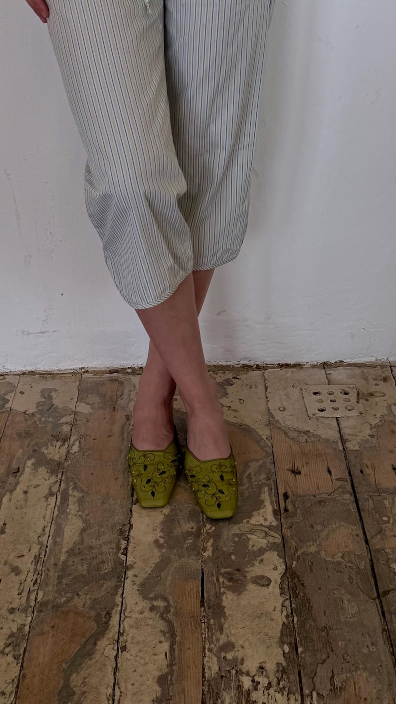 May include: A pair of olive green, embellished mules with a square toe. The shoes are adorned with dark green floral designs and small, round embellishments. The person is wearing striped pants with blue and white stripes.