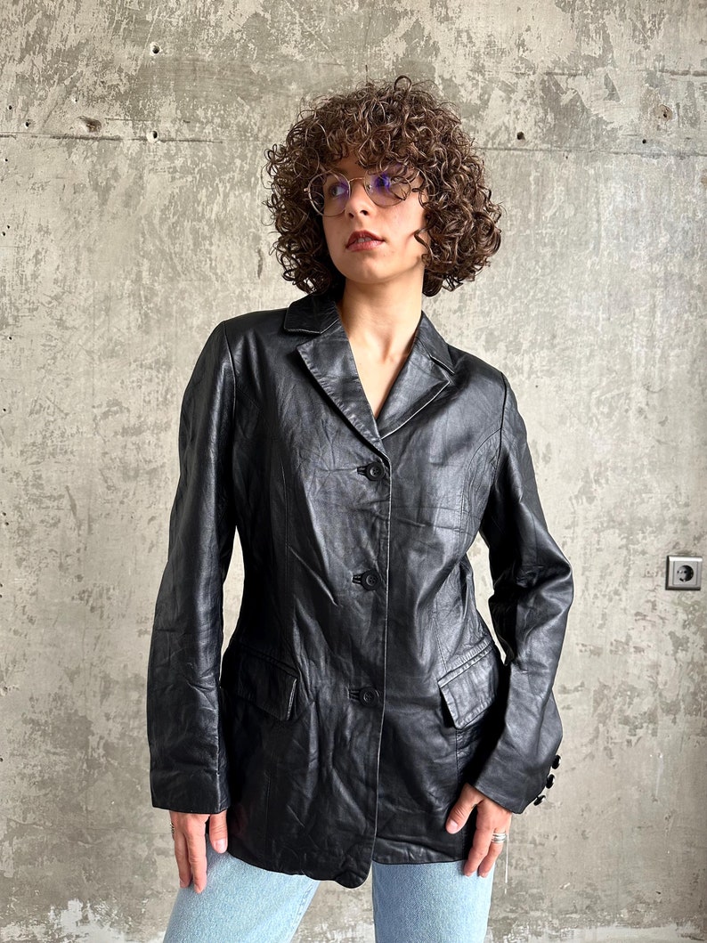 Puede incluir: Un blazer de cuero negro con solapas, cierre de tres botones y dos bolsillos delanteros. El blazer tiene un dise&ntilde;o cl&aacute;sico y un aspecto ligeramente desgastado. La chaqueta la lleva una persona.