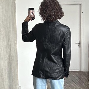 Puede incluir: Una blazer de cuero negro con un dise&ntilde;o cl&aacute;sico. La blazer se lleva con vaqueros azul claro. La persona sostiene un smartphone, tomando una foto frente a una pared blanca y una puerta blanca.