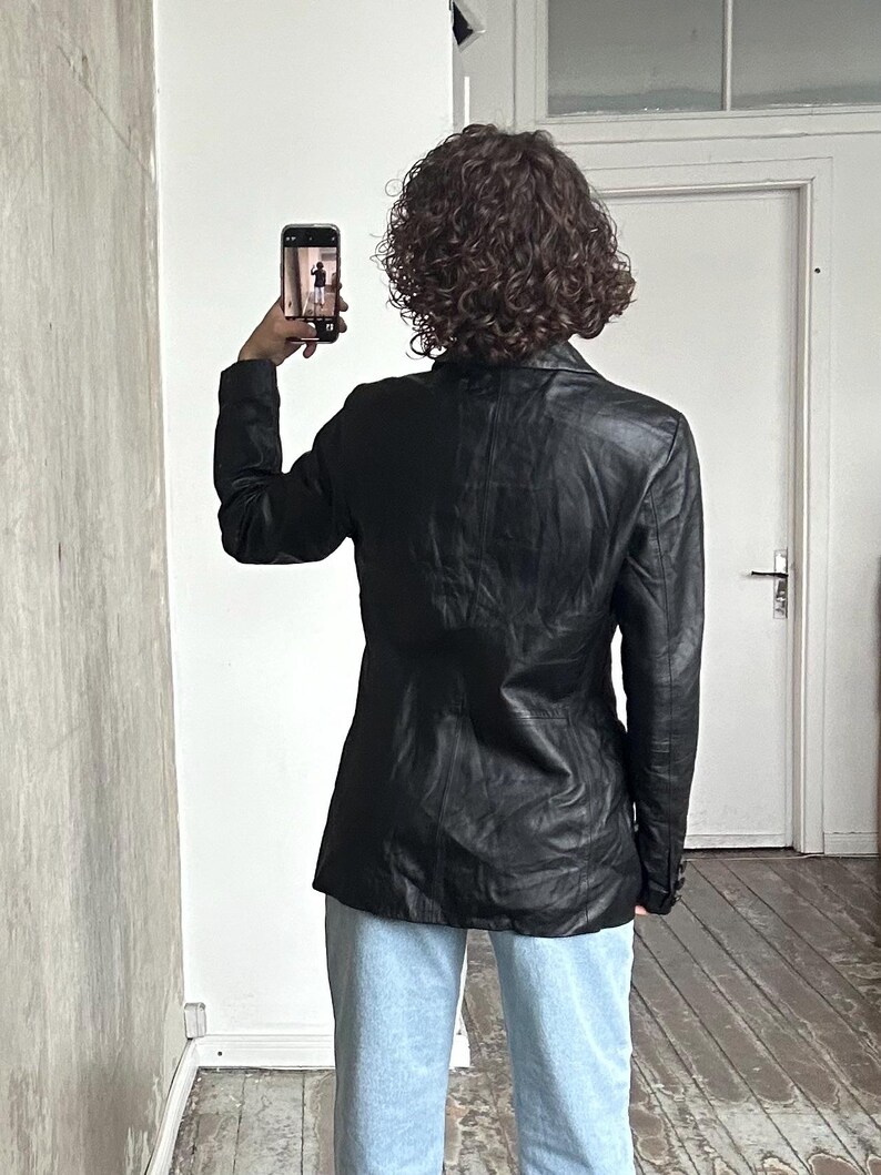 Puede incluir: Una blazer de cuero negro con un dise&ntilde;o cl&aacute;sico. La blazer se lleva con vaqueros azul claro. La persona sostiene un smartphone, tomando una foto frente a una pared blanca y una puerta blanca.