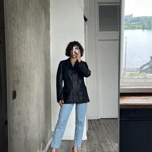 Puede incluir: Un blazer negro con un dise&ntilde;o cl&aacute;sico, combinado con vaqueros azul claro y tacones negros. El blazer tiene un corte entallado y cierre de botones. El atuendo se captura en una habitaci&oacute;n con una ventana que da a un cuerpo de agua.