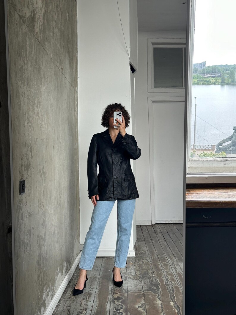 Puede incluir: Un blazer negro con un dise&ntilde;o cl&aacute;sico, combinado con vaqueros azul claro y tacones negros. El blazer tiene un corte entallado y cierre de botones. El atuendo se captura en una habitaci&oacute;n con una ventana que da a un cuerpo de agua.