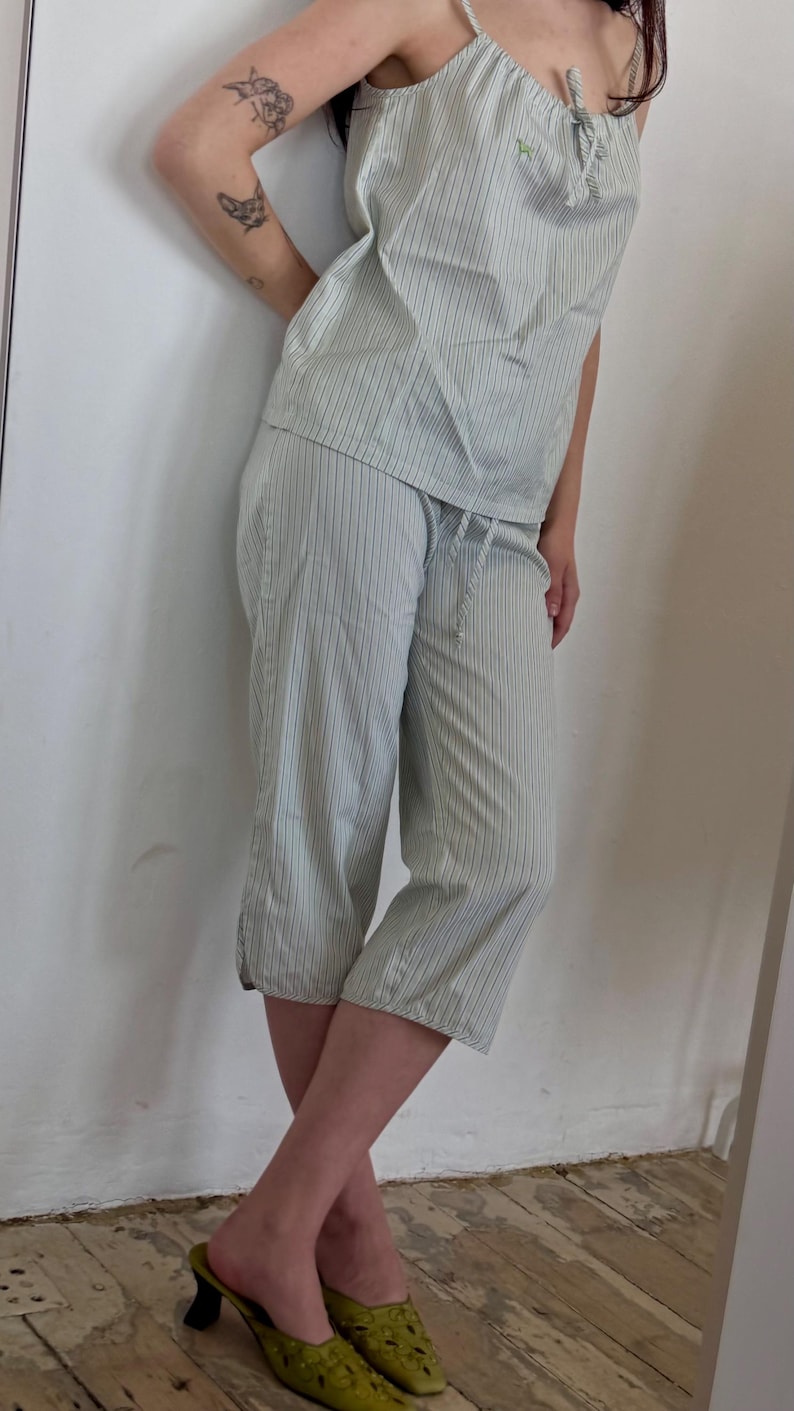 May include: Light green and white striped pajama set. The set includes a camisole top with a bow detail and capri-length pants. The top has thin straps and a small embroidered detail. The pants have a drawstring waist. The model is wearing green mules.
