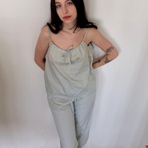 May include: A light green and white striped pajama set. The set includes a camisole top with thin straps and a drawstring pajama pant. The model is wearing green shoes. The background is a white wall and a wooden floor.