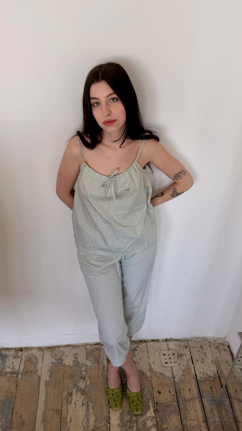 May include: A light green and white striped pajama set. The set includes a camisole top with thin straps and a drawstring pajama pant. The model is wearing green shoes. The background is a white wall and a wooden floor.