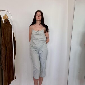 May include: A light blue and white striped pajama set. The set includes a camisole top with thin straps and matching capri pants. The person is wearing green patterned shoes. A clothing rack and mirror are in the background.