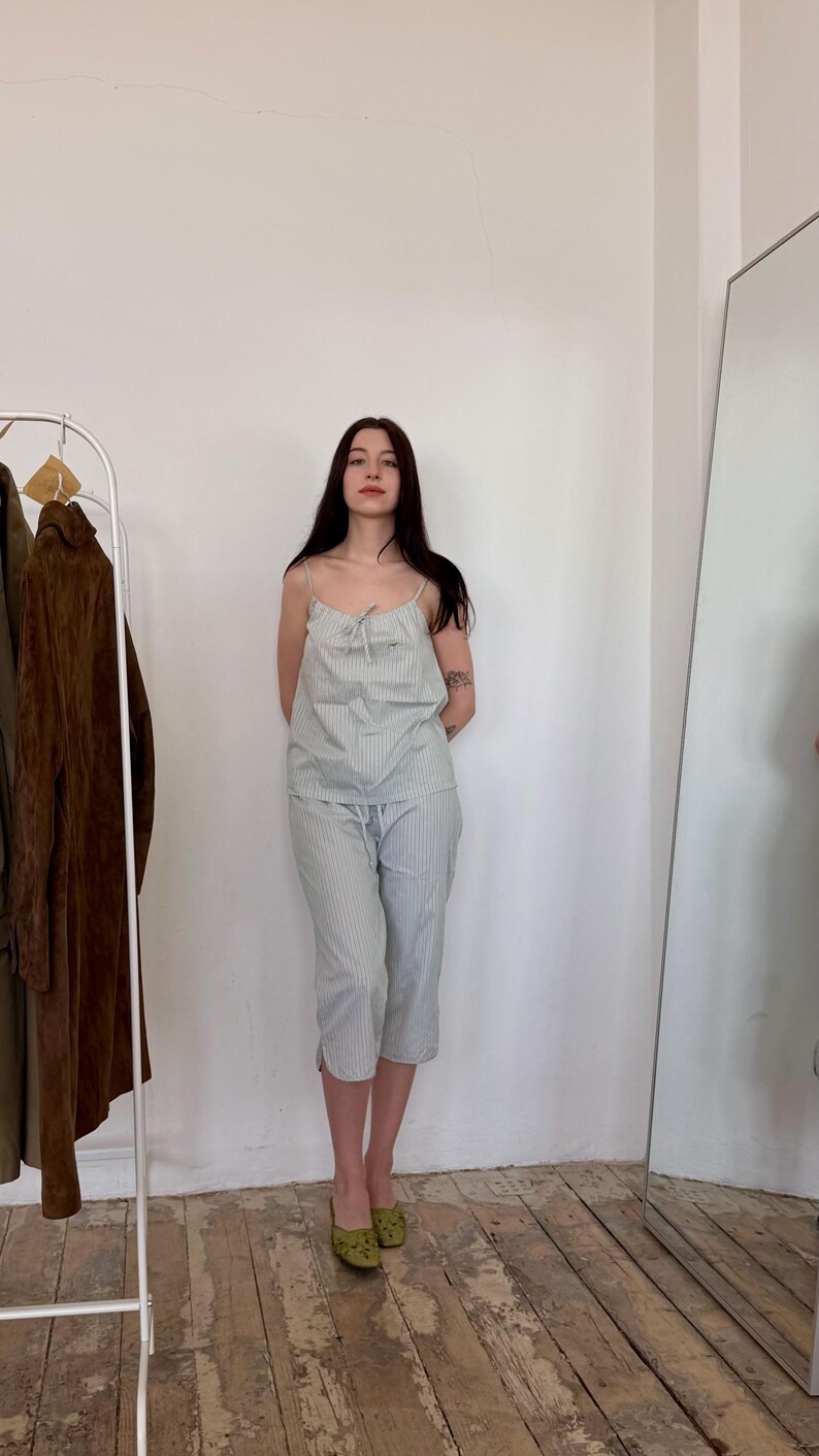 May include: A light blue and white striped pajama set. The set includes a camisole top with thin straps and matching capri pants. The person is wearing green patterned shoes. A clothing rack and mirror are in the background.