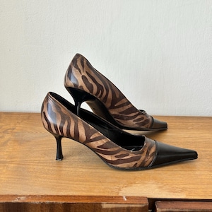 Könnte beinhalten: Ein Paar Pumps mit Zebradruck, schwarzen spitzen Zehen und Absätzen. Die Absätze sind etwa 7,6 cm hoch. Die Schuhe sind aus einem braun-schwarz gemusterten Material.
