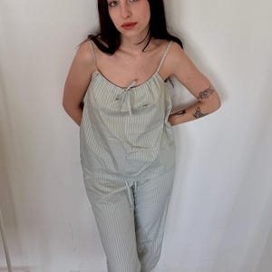 May include: A light green and white striped pajama set. The set includes a camisole top with thin straps and a drawstring waist pant. The model is wearing green patterned shoes. The background is a white wall and a wooden floor.