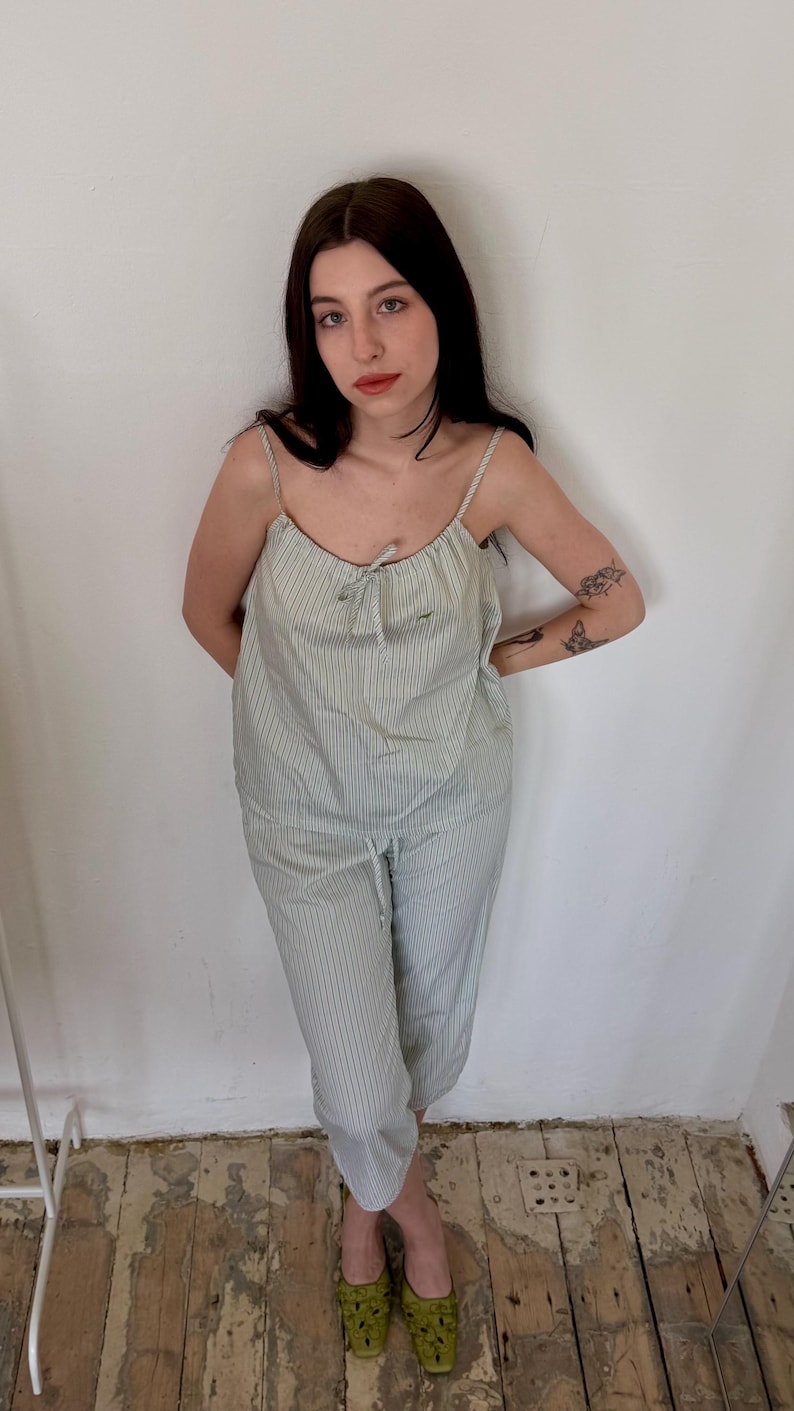 May include: A light green and white striped pajama set. The set includes a camisole top with thin straps and a drawstring waist pant. The model is wearing green patterned shoes. The background is a white wall and a wooden floor.