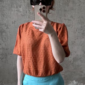 Puede incluir: Una persona con una camiseta naranja texturizada de manga corta y una falda azul claro. La camiseta tiene un patrón sutil y un escote redondo. La persona lleva gafas de sol negras y sostiene un smartphone.