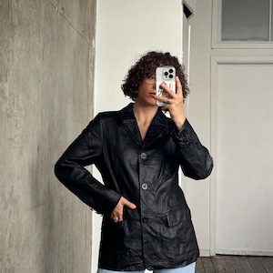 Puede incluir: Un blazer de cuero negro con solapas, cierre de tres botones y dos bolsillos delanteros. El blazer se lleva con vaqueros azul claro. La chaqueta tiene un dise&ntilde;o cl&aacute;sico y entallado.