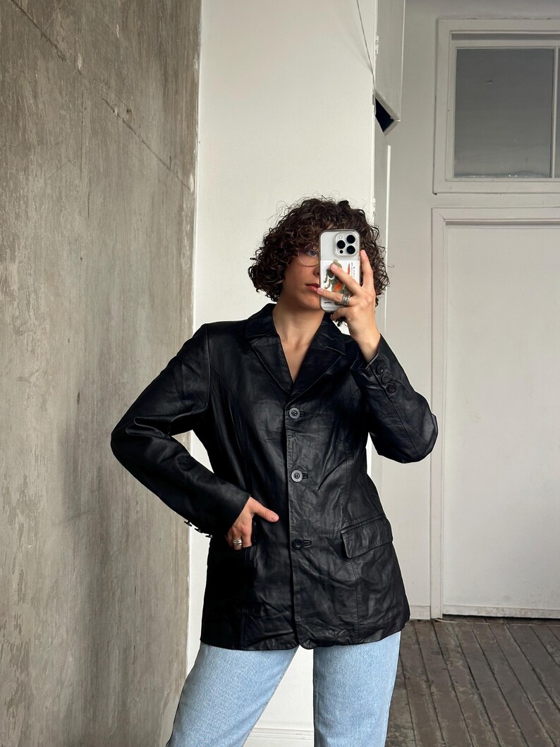 Puede incluir: Un blazer de cuero negro con solapas, cierre de tres botones y dos bolsillos delanteros. El blazer se lleva con vaqueros azul claro. La chaqueta tiene un dise&ntilde;o cl&aacute;sico y entallado.