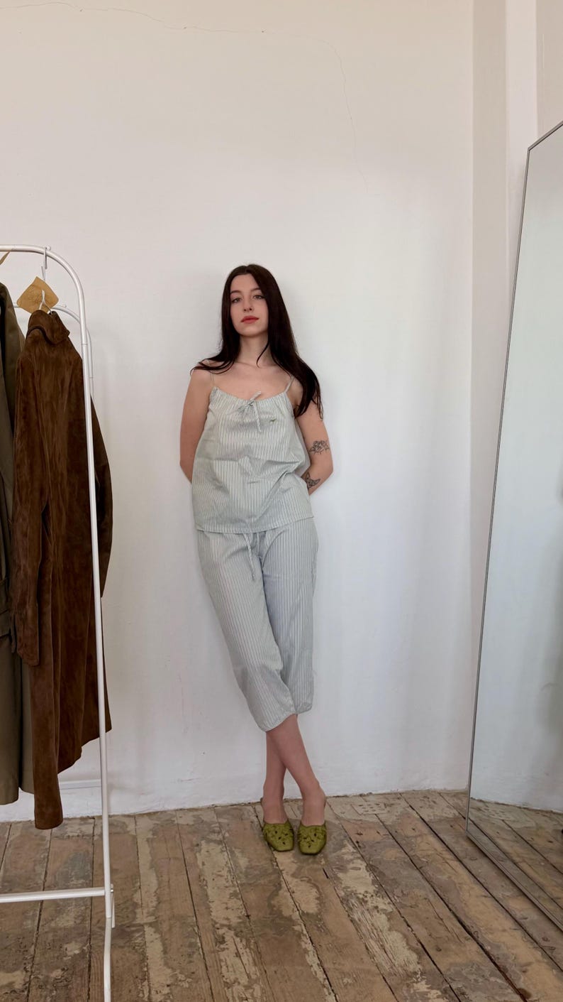 May include: A light blue and white striped pajama set. The set includes a camisole top with a bow detail and matching cropped pants. The person is wearing green patterned shoes. A clothing rack and mirror are in the background.