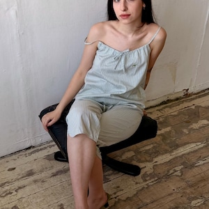 May include: A light blue and white striped pajama set. The set includes a camisole top with a bow and matching capri pants. The person is wearing green slip-on shoes. The person is sitting on a black chair.