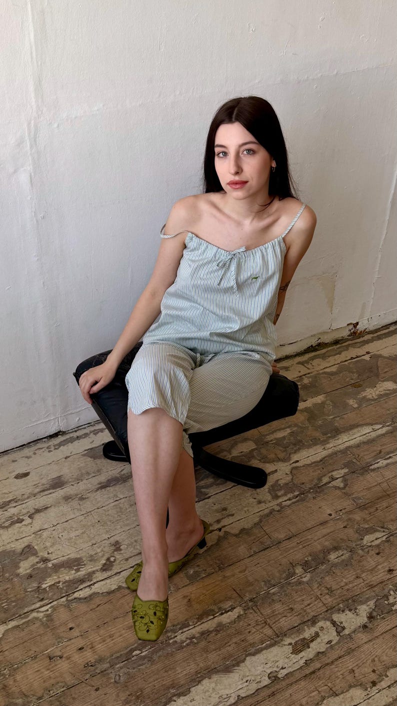 May include: A light blue and white striped pajama set. The set includes a camisole top with a bow and matching capri pants. The person is wearing green slip-on shoes. The person is sitting on a black chair.